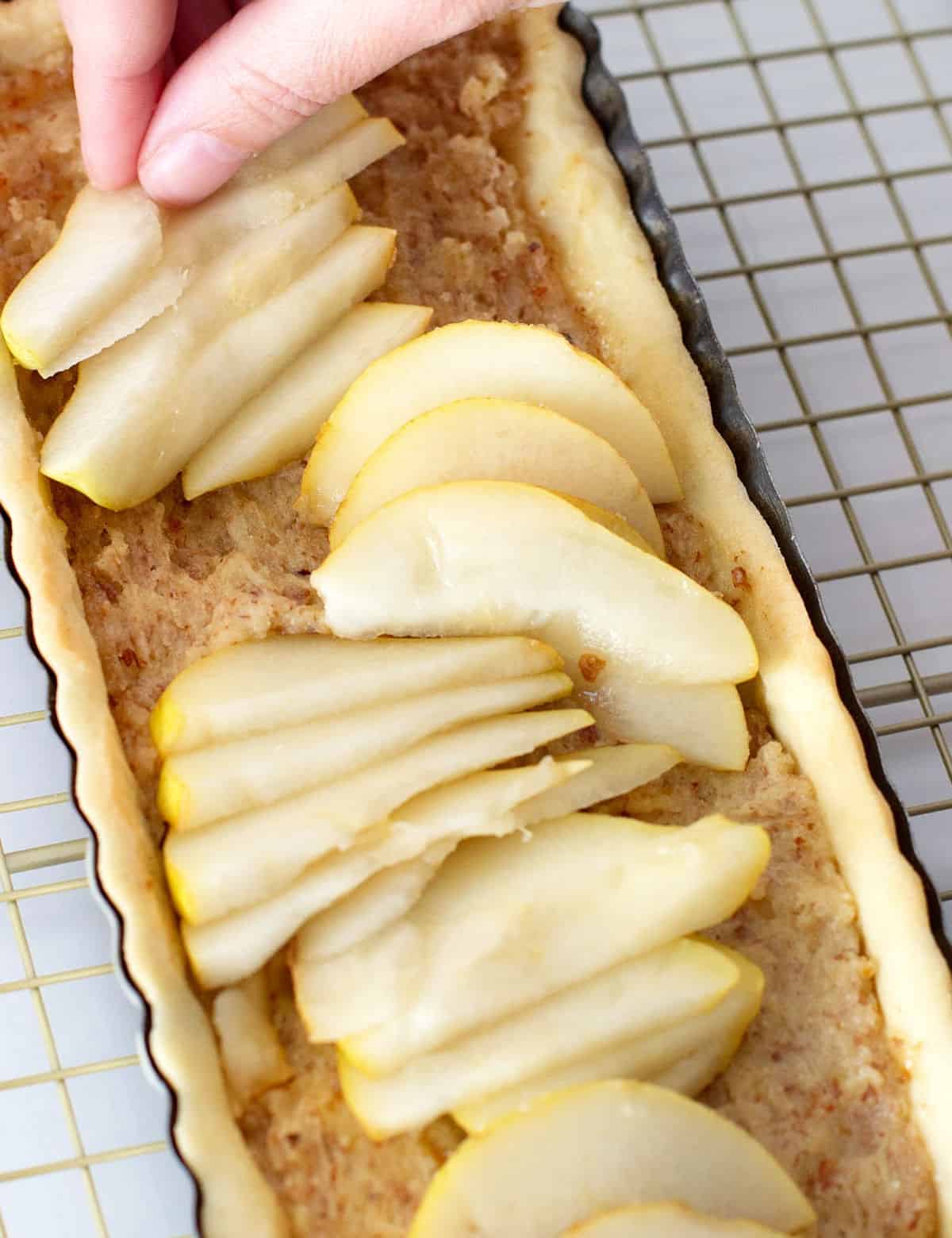 Adding pear slices by hand to long almond tart on a golden wire rack.
