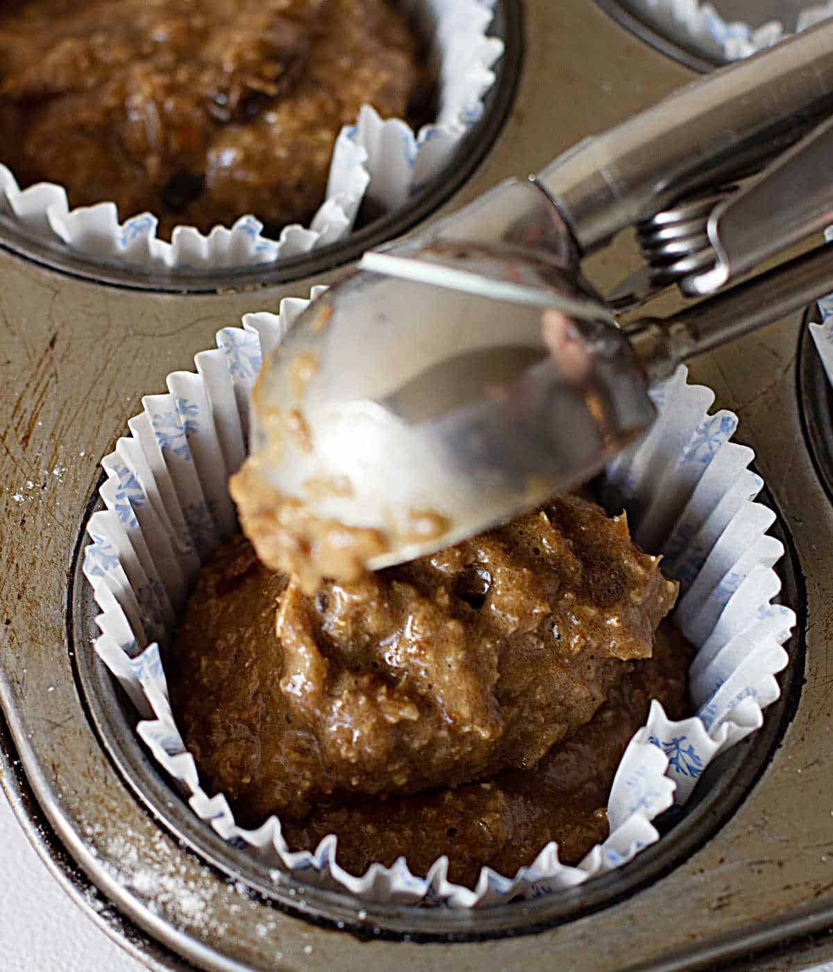 Scooping bran muffin batter in paper liners on a metal muffin tin.