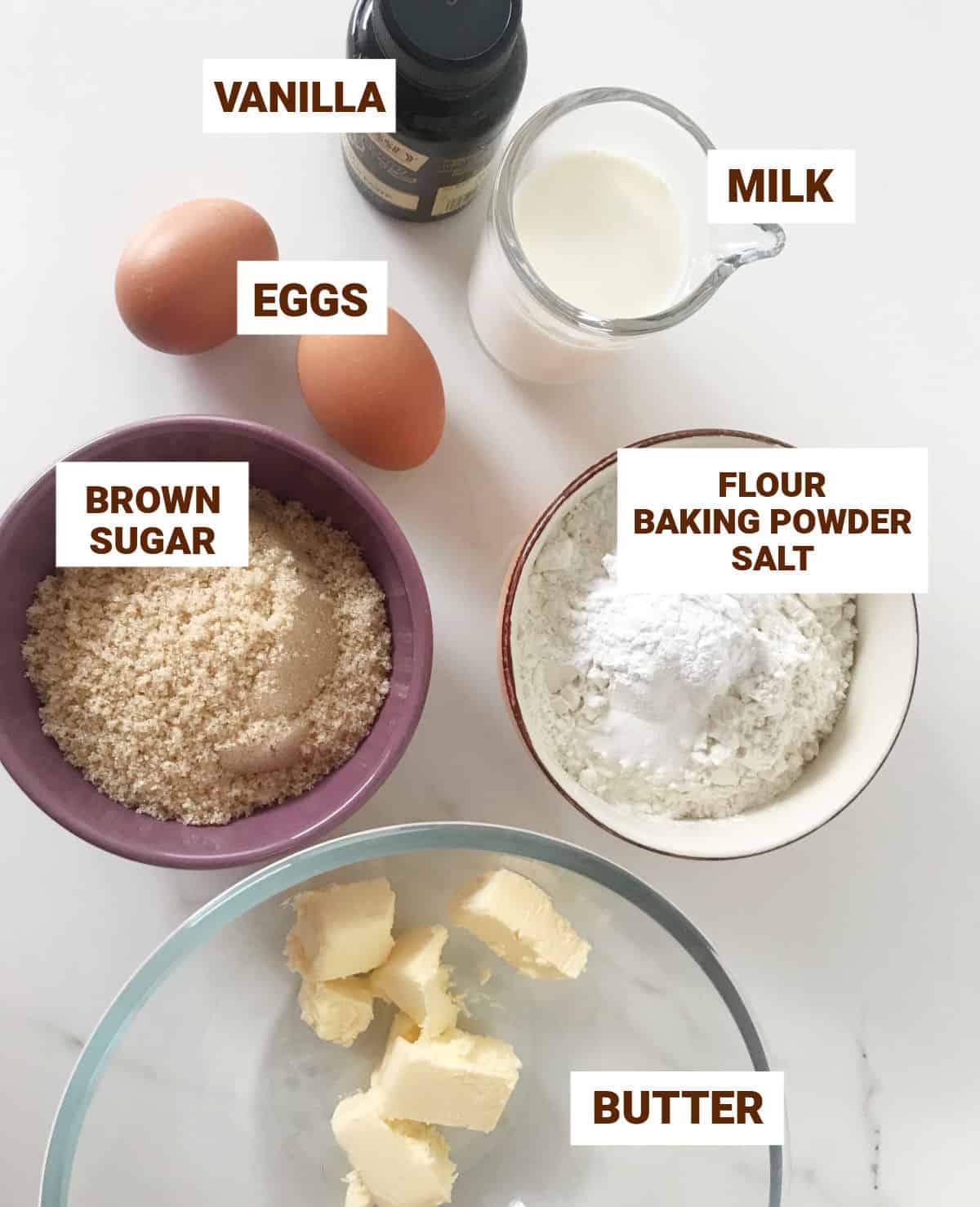 White surface with bowls containing ingredients for brown sugar cake including butter, eggs, vanilla, flour, milk.