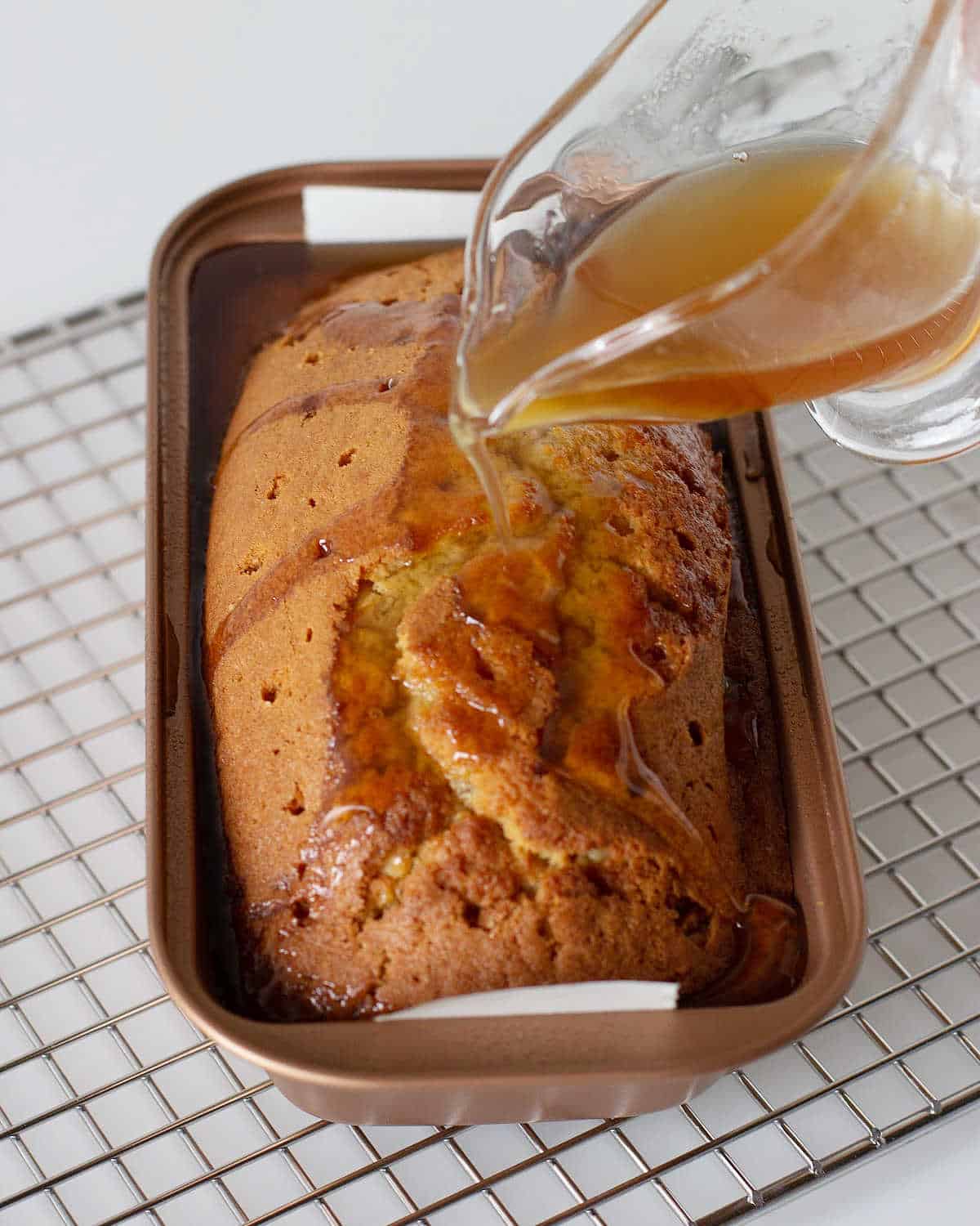 Pouring syrup from a glass jar on top of a brown sugar loaf in the pan on a wire rack.