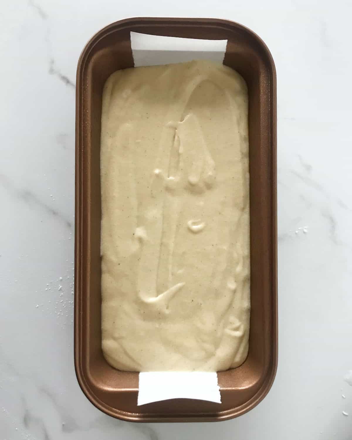 Metal loaf pan with ginger cake batter on a white marble surface.