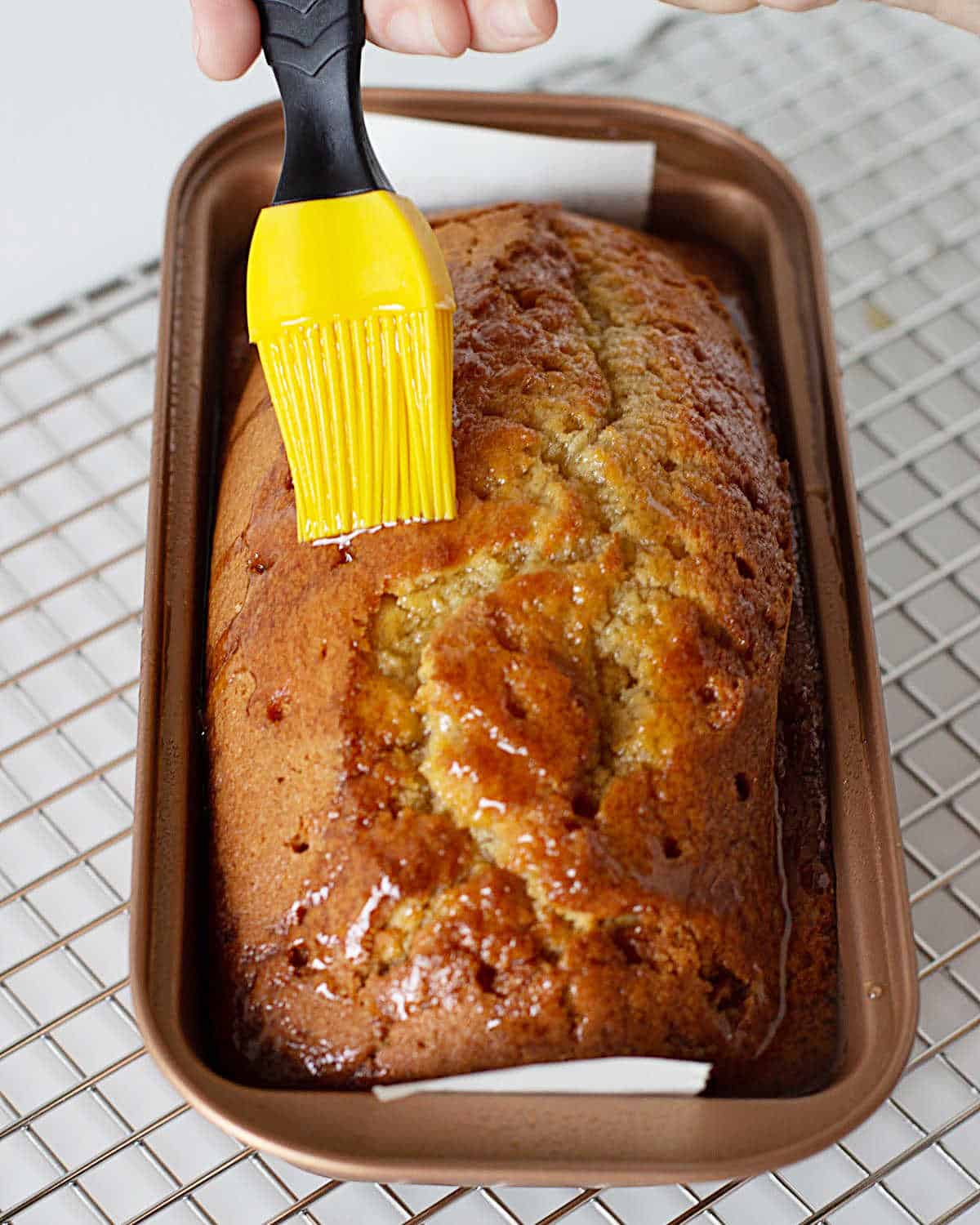 Brushing syrup with a yellow brush on top of a brown sugar loaf in the pan on a wire rack.