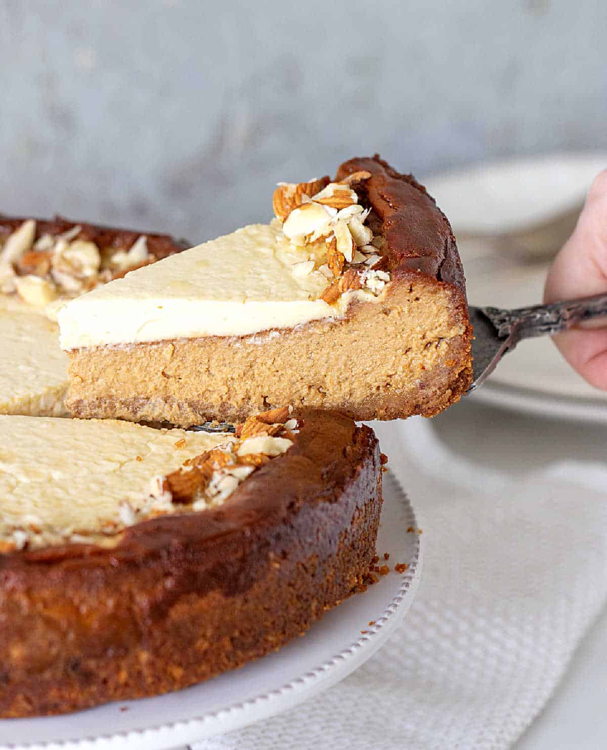 Lifting a slice of brown sugar cheesecake from cake on a white cake stand. Grey background.