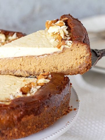 Hand lifting slice of brown sugar cheesecake with a cake server from whole cheesecake on a white plate. Grey background.