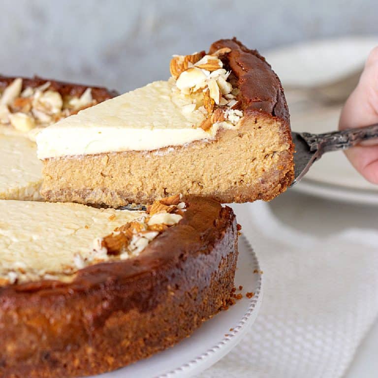 Hand lifting slice of brown sugar cheesecake with a cake server from whole cheesecake on a white plate. Grey background.