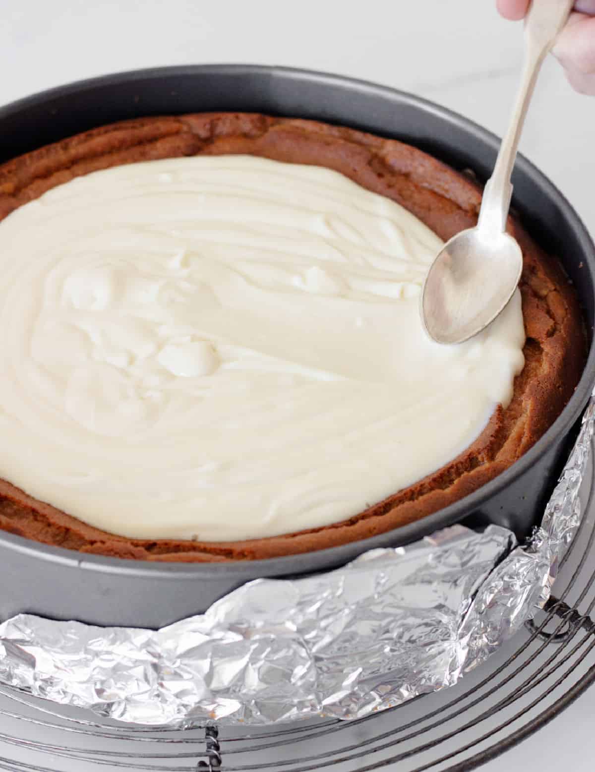 Spreading sour cream topping with a silver spoon on top of brown sugar cheesecake in the pan.