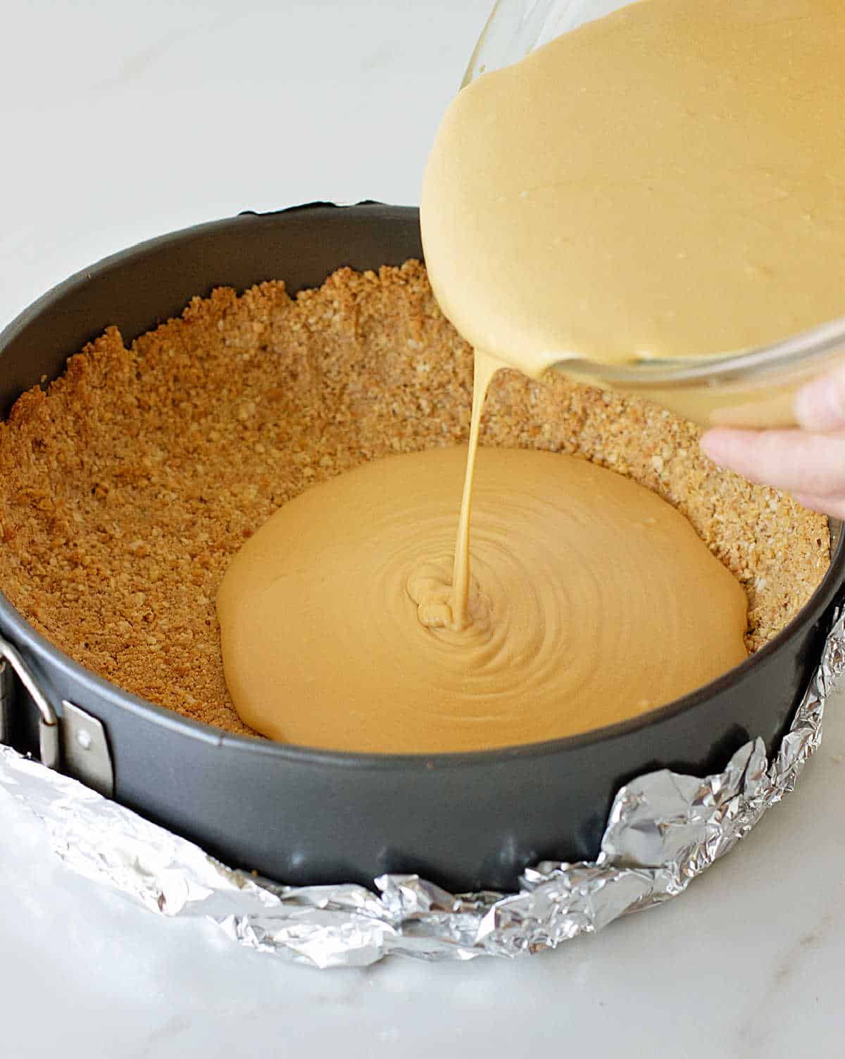 Pouring brown sugar cheesecake filling into crumb crust on a round cake pan with aluminum foil in the bottom. White surface and background.