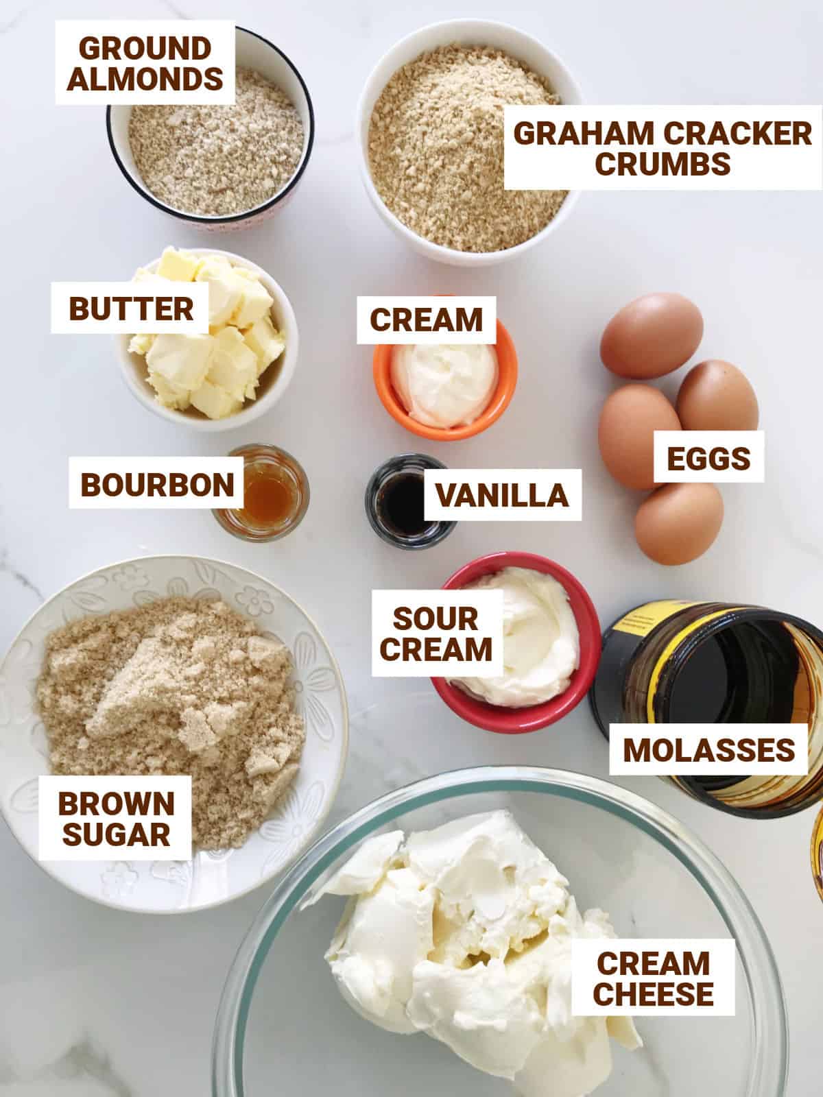 Brown sugar cheesecake ingredients in bowls on white surface including molasses, butter, almonds, cream, graham crackers, eggs, flavorings.