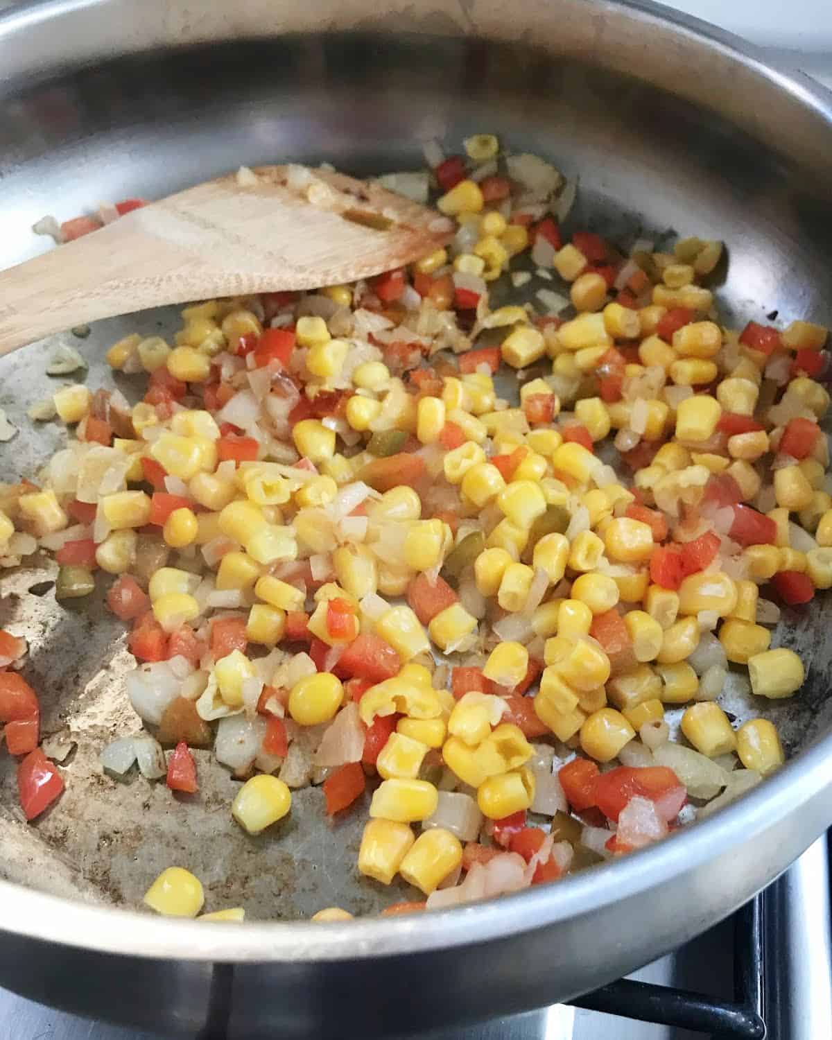 Corn, chopped onions and jalapeños being sauteed in a metal saucepan with a wooden spoon.