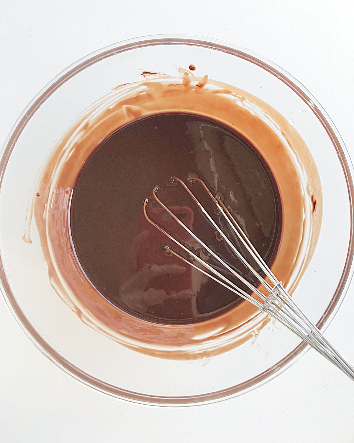A whisk inside a glass bowl with melted chocolate and butter on a white surface.