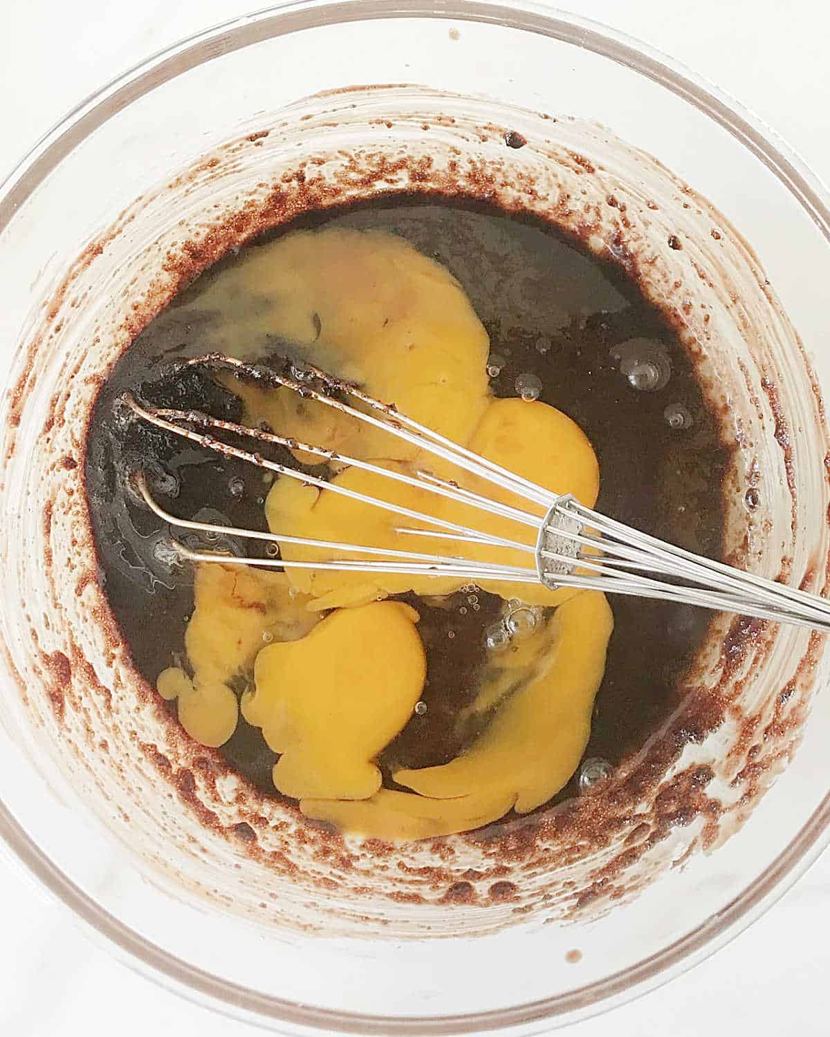 Eggs and melted chocolate in a glass bowl with a whisk. White surface. 
