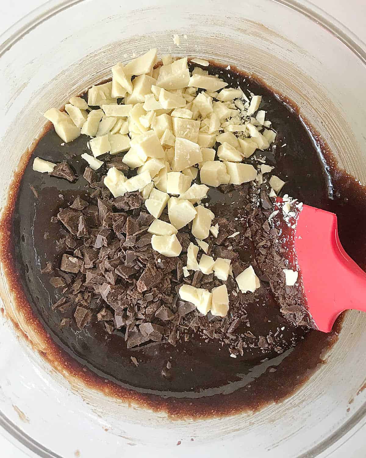 White and dark chocolate chunks added to brownie batter in a glass bowl. Red spatula. White surface.