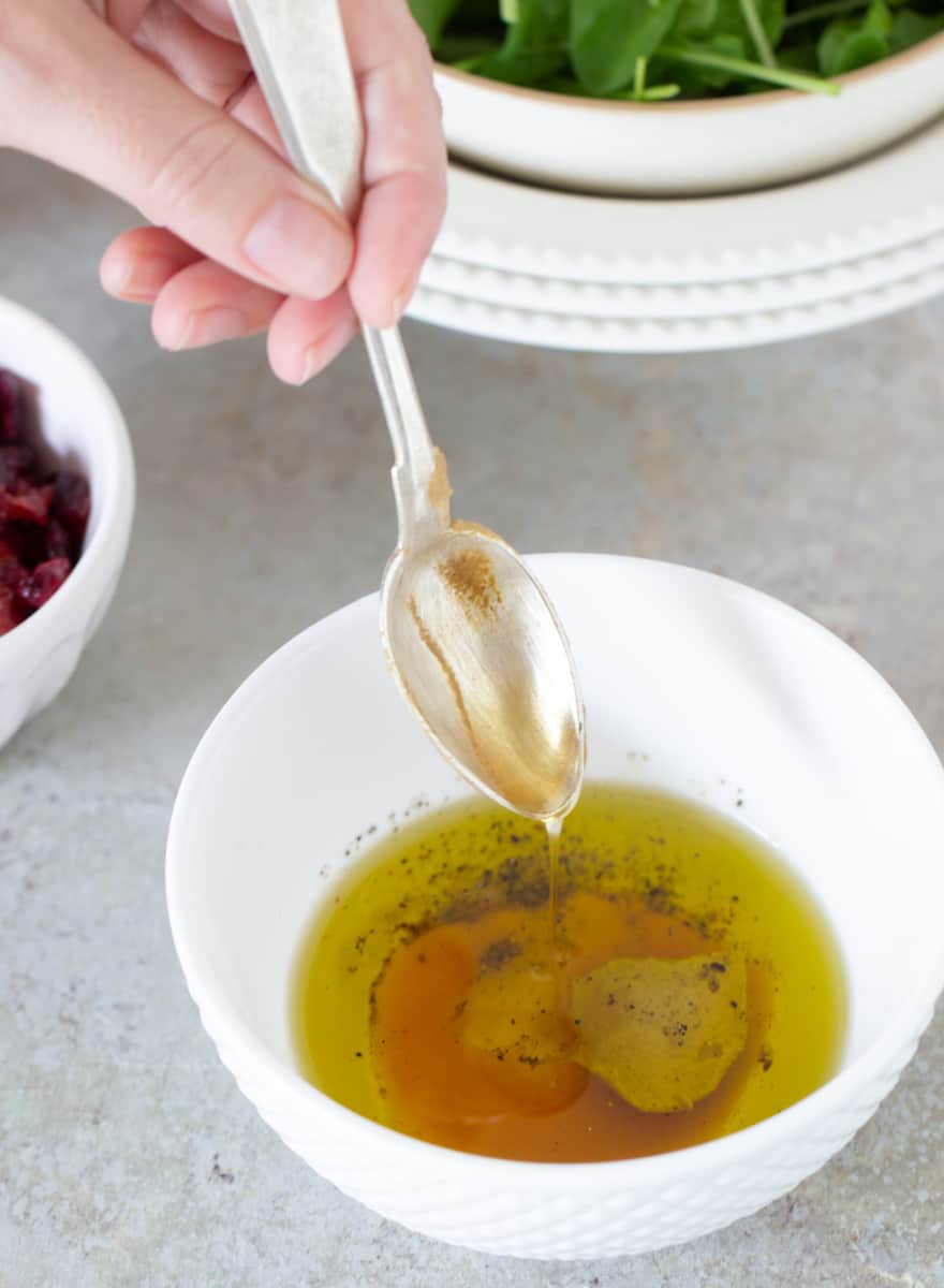 Adding honey with spoon to white bowl with vinaigrette.
