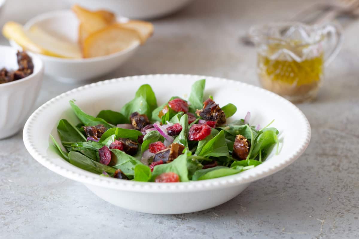 Arugula and dried fruit in white bowl plate, bowls, vinagrette, grey background.