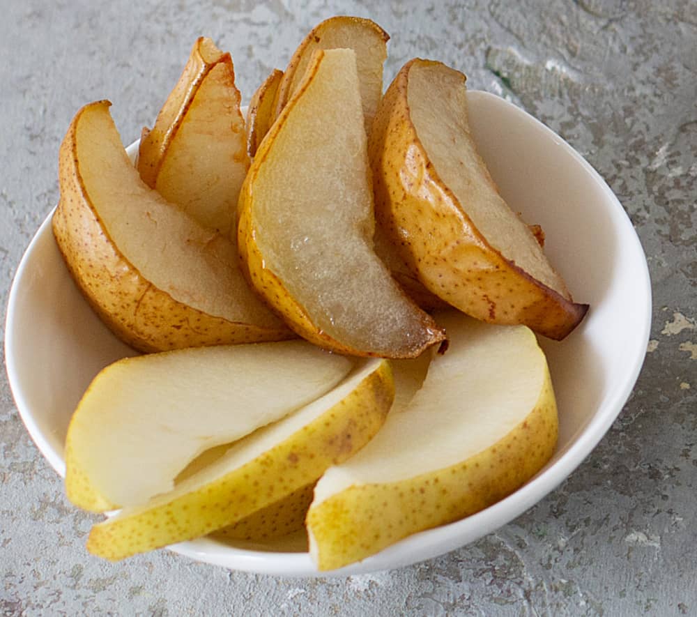 Raw and baked pear wedges on white bowl, grey surface.