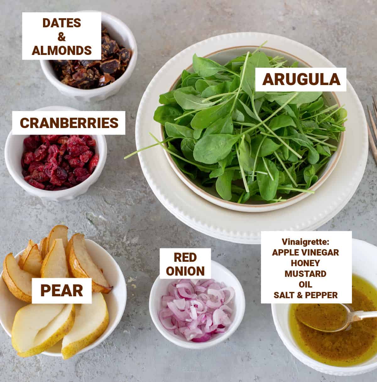 Pear arugula salad ingredients in bowls on grey surface including dates, onion, vinaigrette, cranberries, almonds.