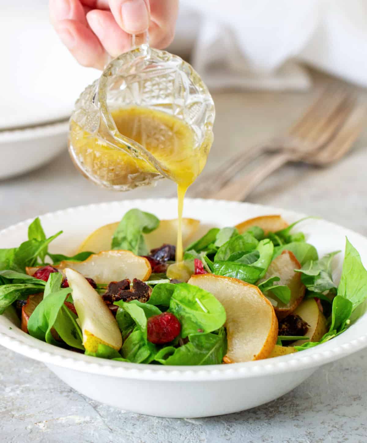 Pouring thick vinaigrette over white plate with arugula and pear salad. Grey surface. 