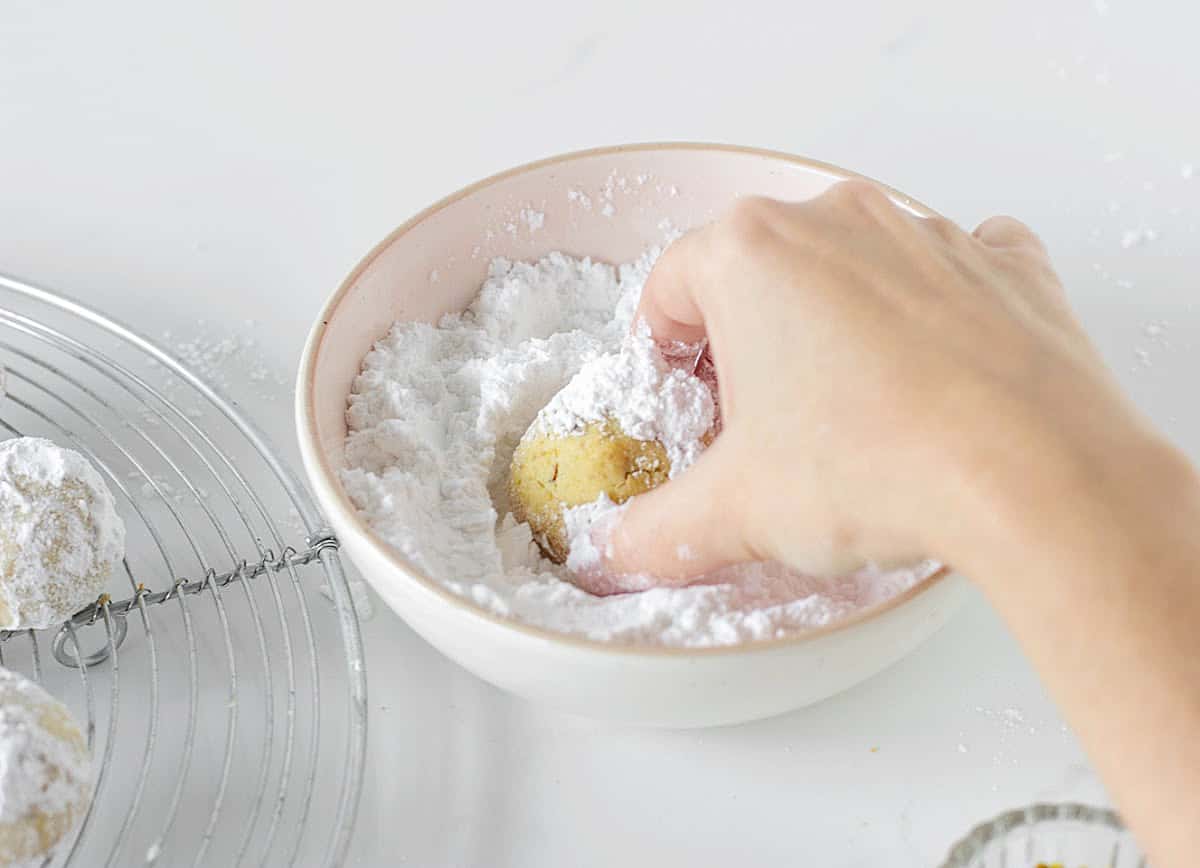 Hand rolling pistachio cookie round in a pink bowl with powdered sugar.