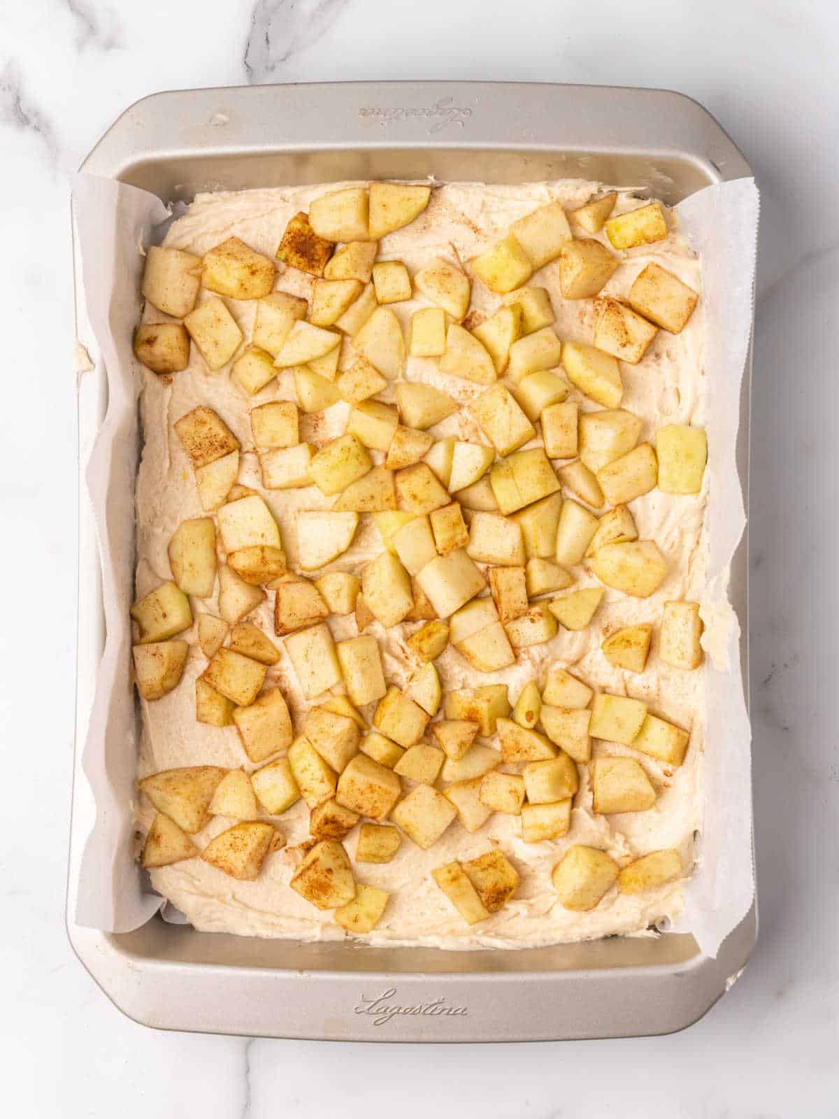 Cake batter and diced apples on a rectangular metal pan placed on white marble surface.