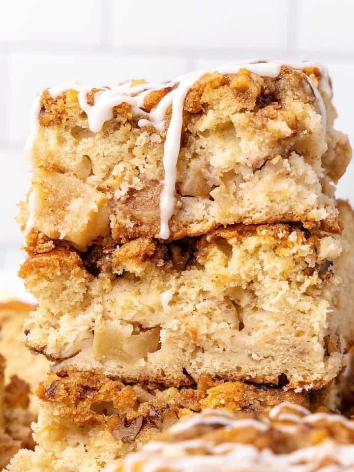 Close up of apple coffee cake squares in a stack with white tile background. Top one is dripping glaze.