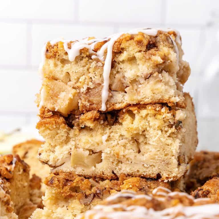 Close up of apple coffee cake squares in a stack with white tile background. Top one is dripping glaze.