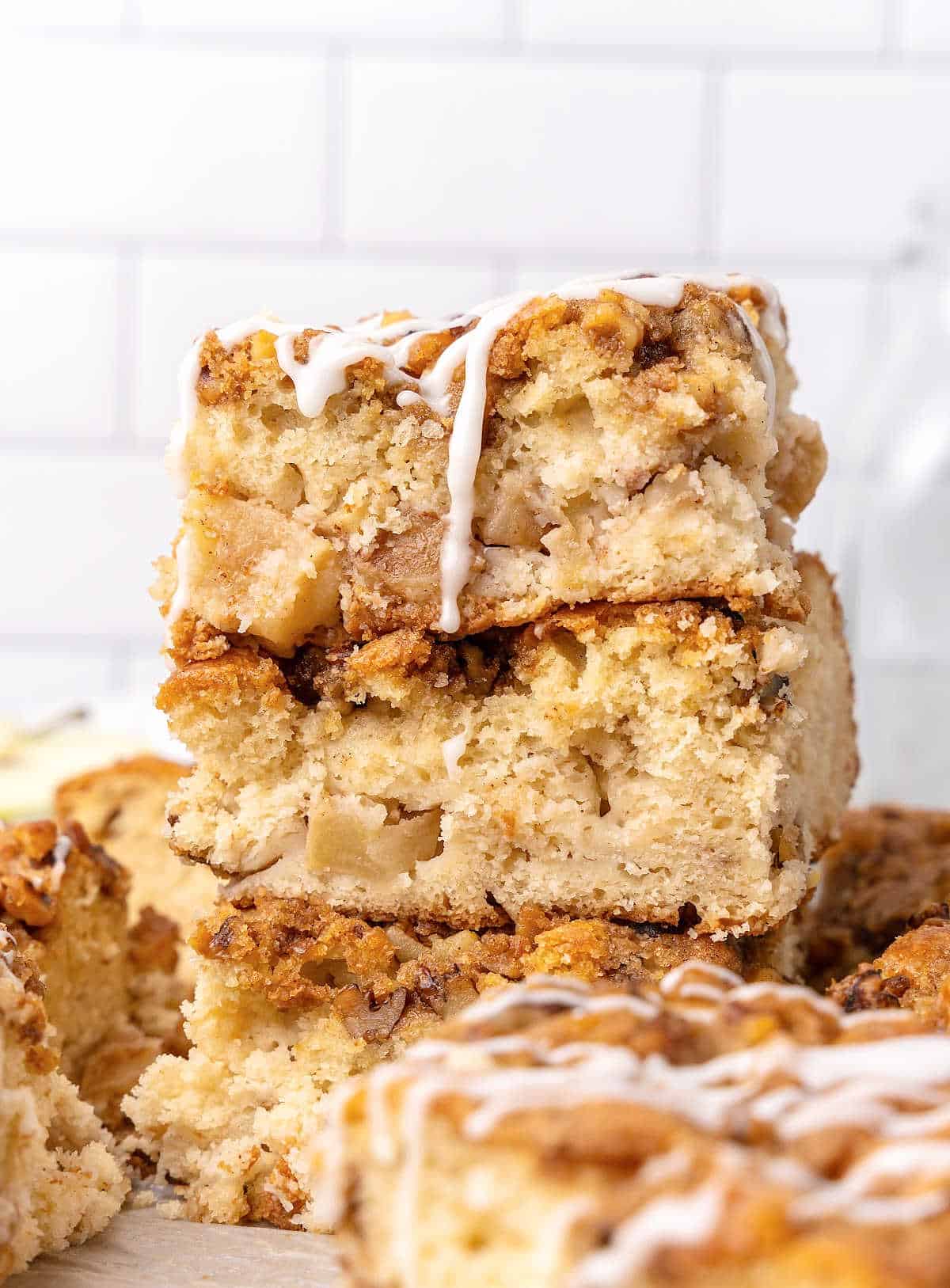 Stack of apple coffee cake squares with glaze. White tile background. 