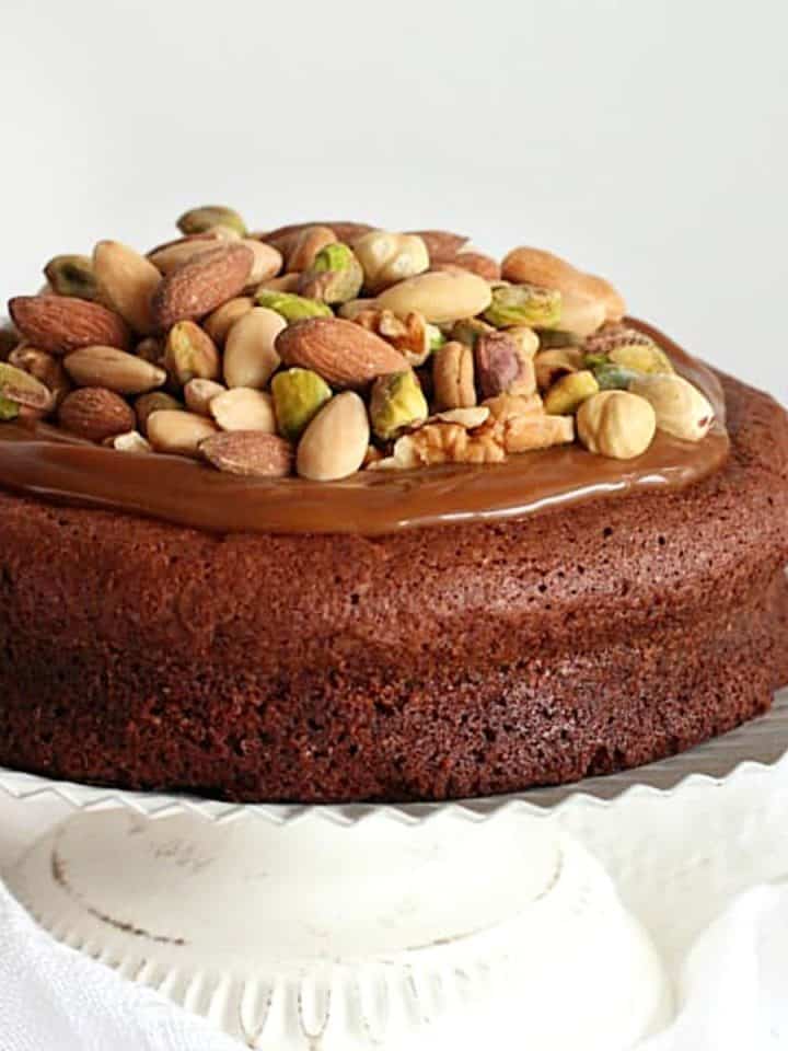 Caramel and nut topped chocolate cake on a white cake stand with white background.
