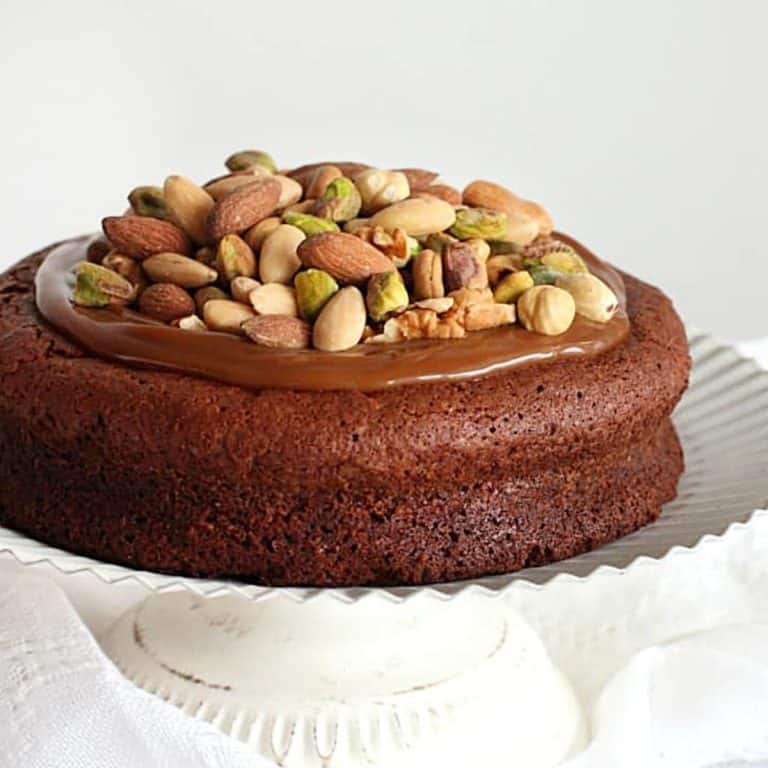 Caramel and nut topped chocolate cake on a white cake stand with white background.