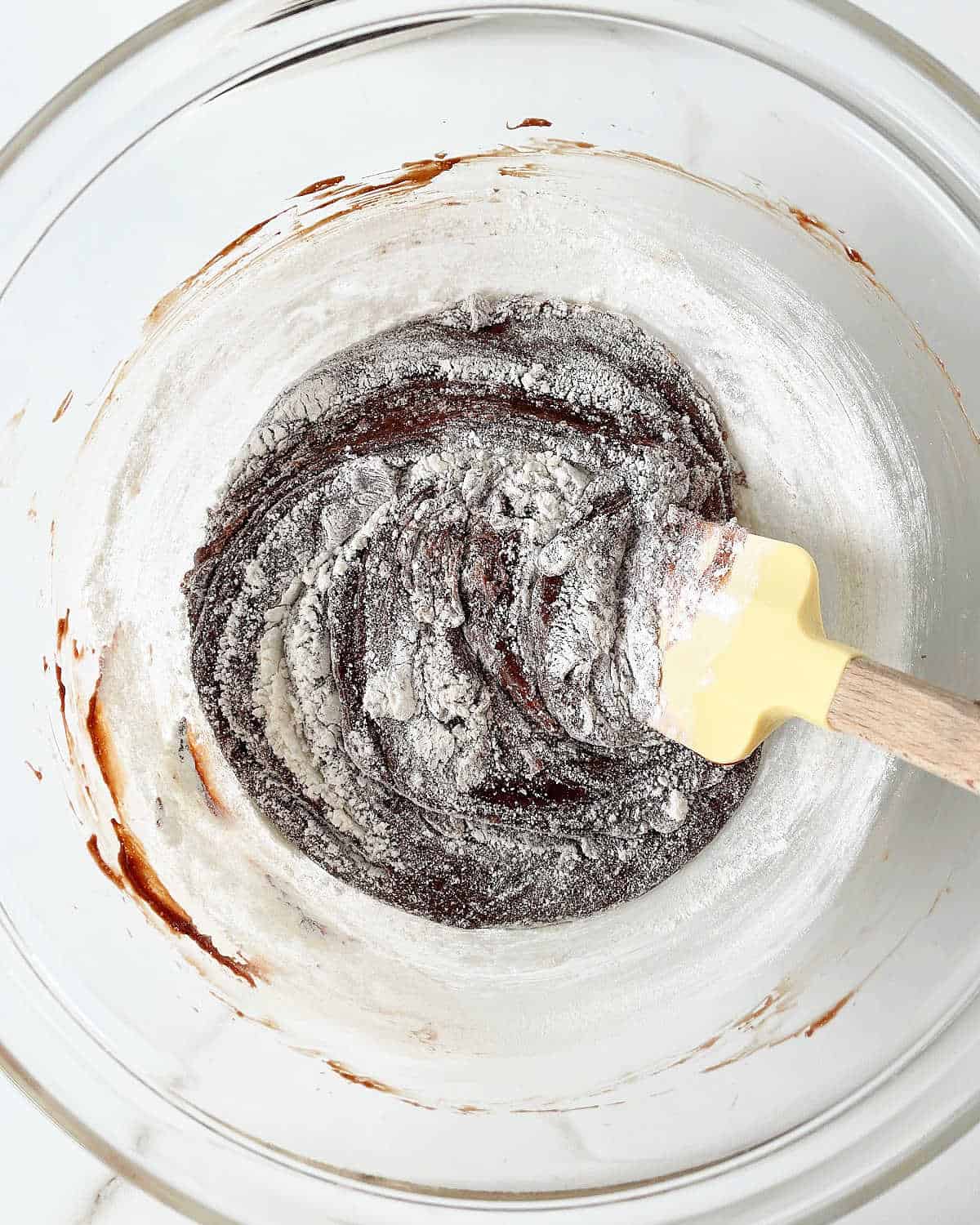 Flour stirred white a yellow spatula into a chocolate batter in a glass bowl.