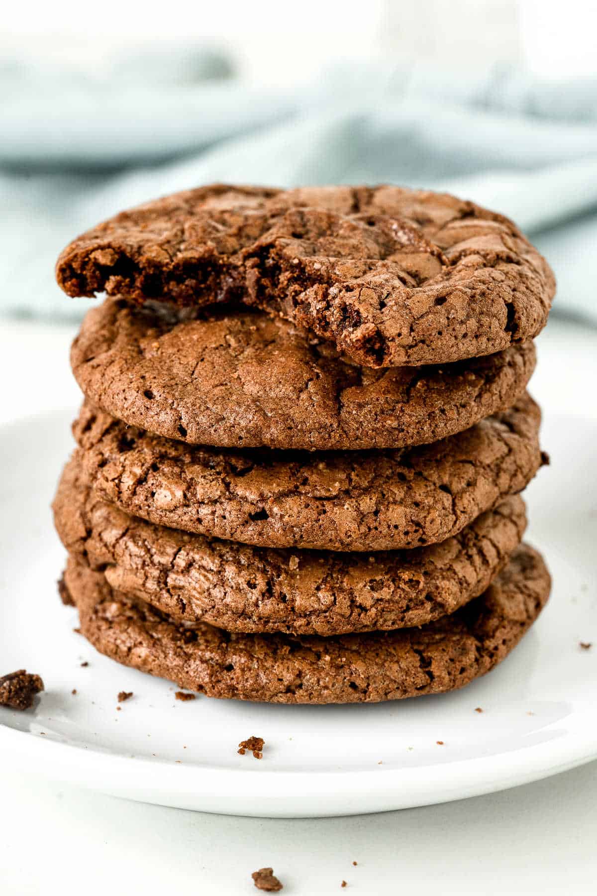 Stack of brownie cookies, the top one is bitten on a white plate. Blue cloth in the background.