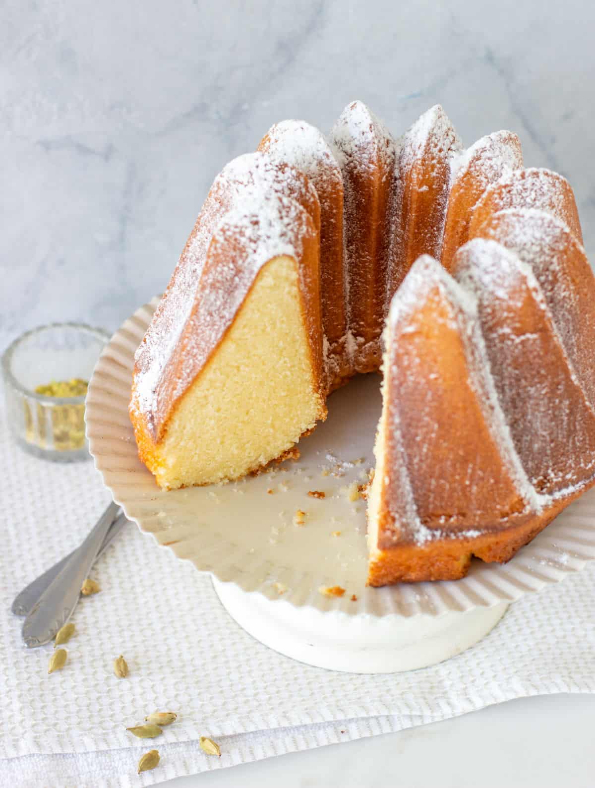 Grey background, white surface with cardamom pods, bundt cake on white cake stand, one piece missing