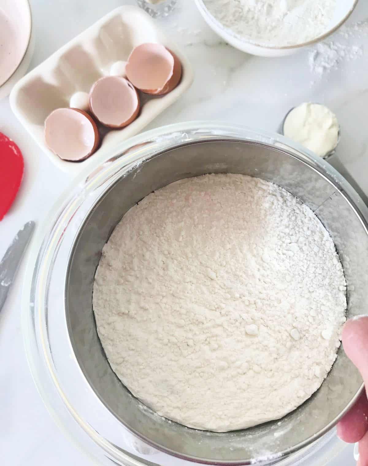 White marble surface with eggs, bowls, hand sifting flour over bowl