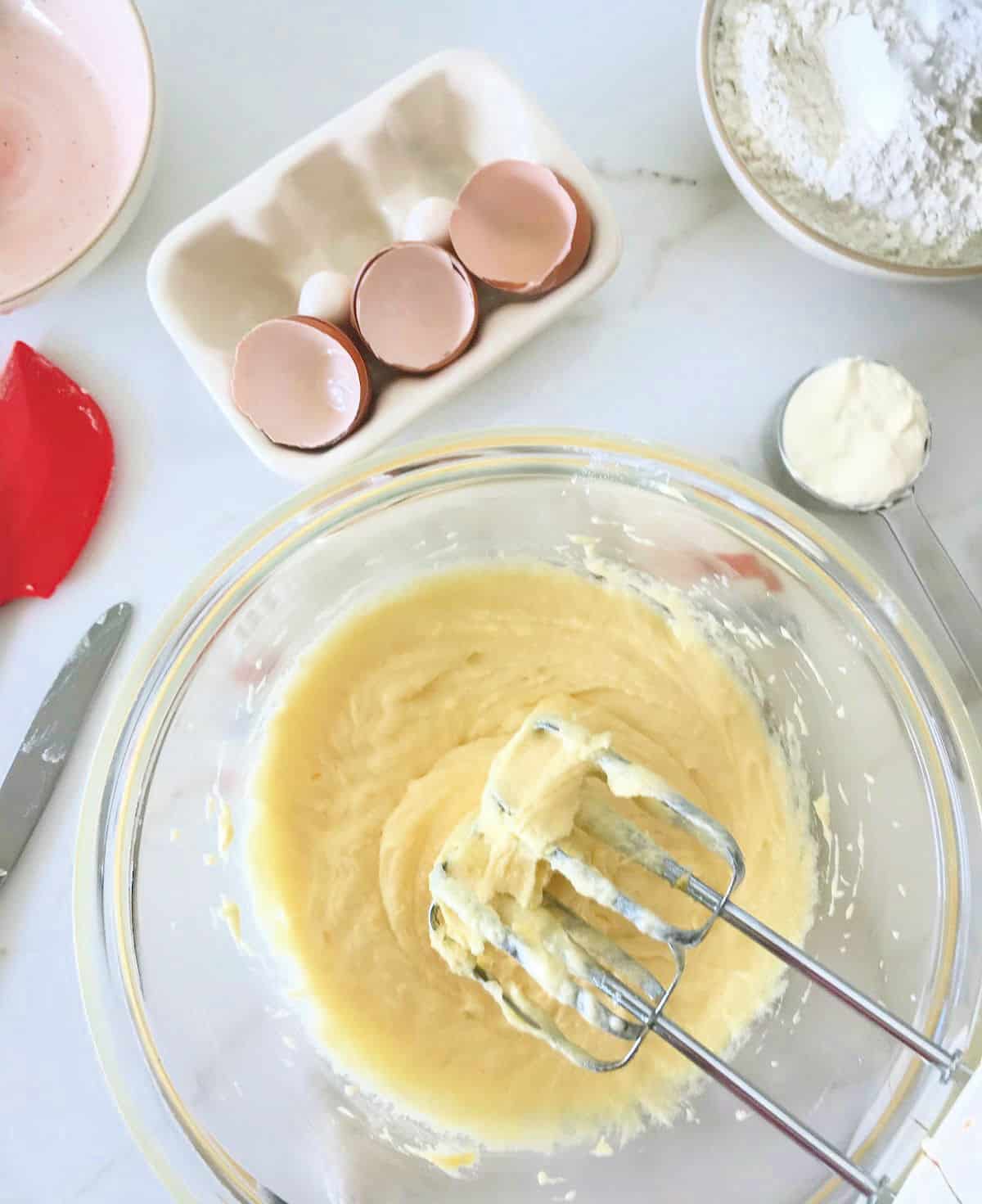 Glass bowl with cake batter, beater, eggs, bowls, white marble surface