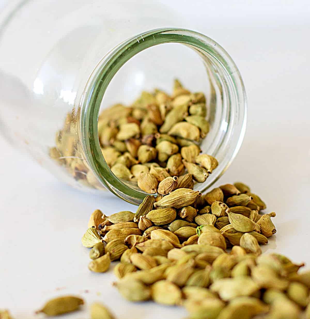 Glass jar with cardamom pods lying on white surface