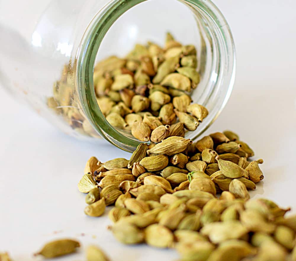 Glass jar with cardamom pods lying on white surface.