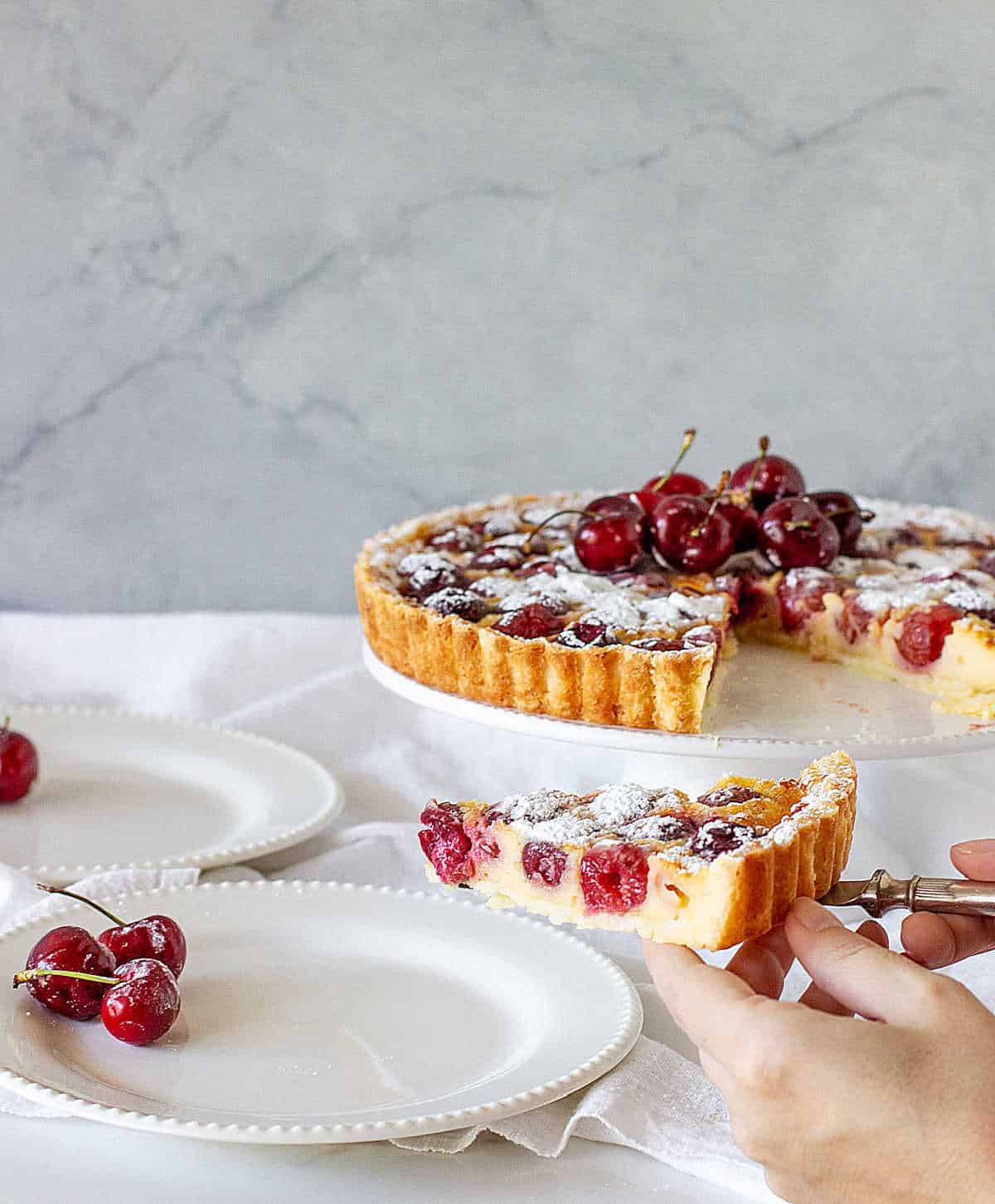 Serving a slice of cherry pie on white plate, tart on cake stand, white background