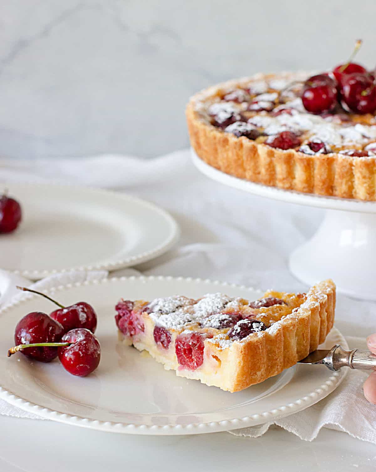 Placing a slice of cherry pie on white plate, tart on cake stand, white background.