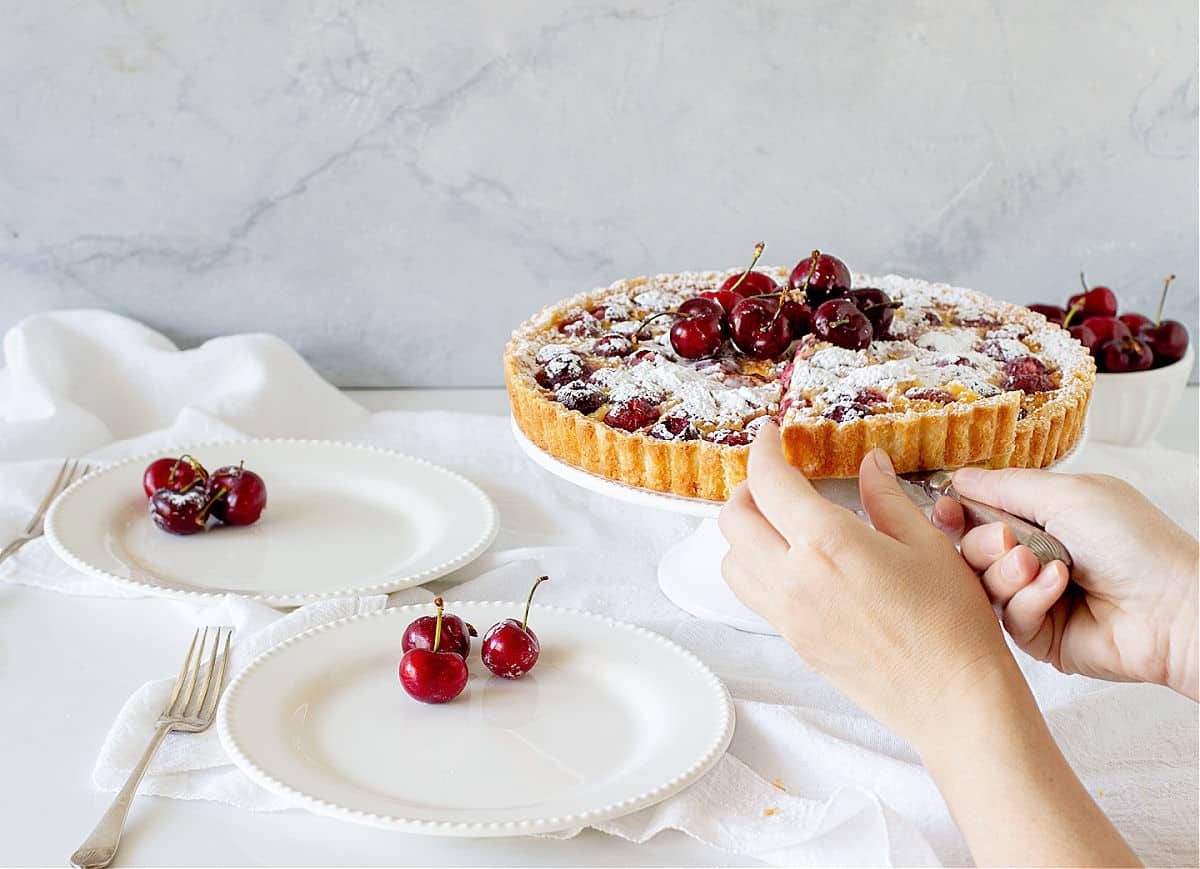 White surface with white plates, cherries, hands cutting whole tart on cake stand.