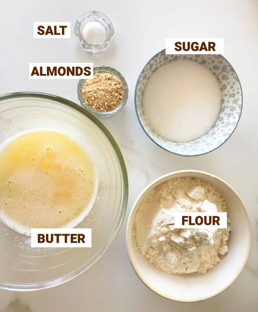 Tart crust ingredients on white marble in bowls including butter, almonds, sugar, salt, flour.