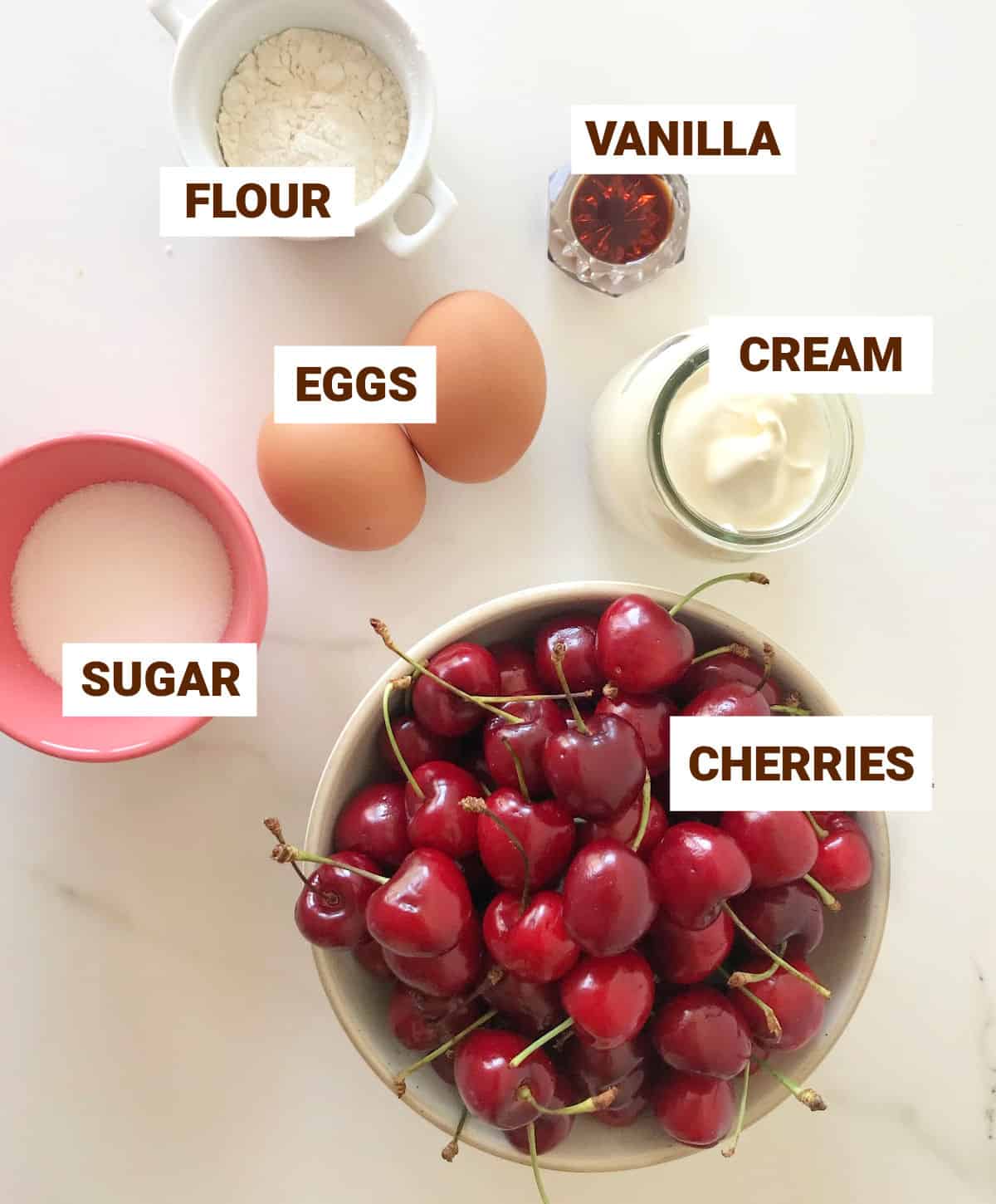White surface with ingredients for cherry tart in bowls including sugar, cream, eggs, vanilla, flour.