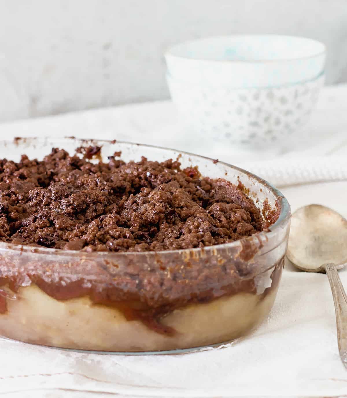 A glass oval dish with baked chocolate apple crisp, grey background, a silver spoon