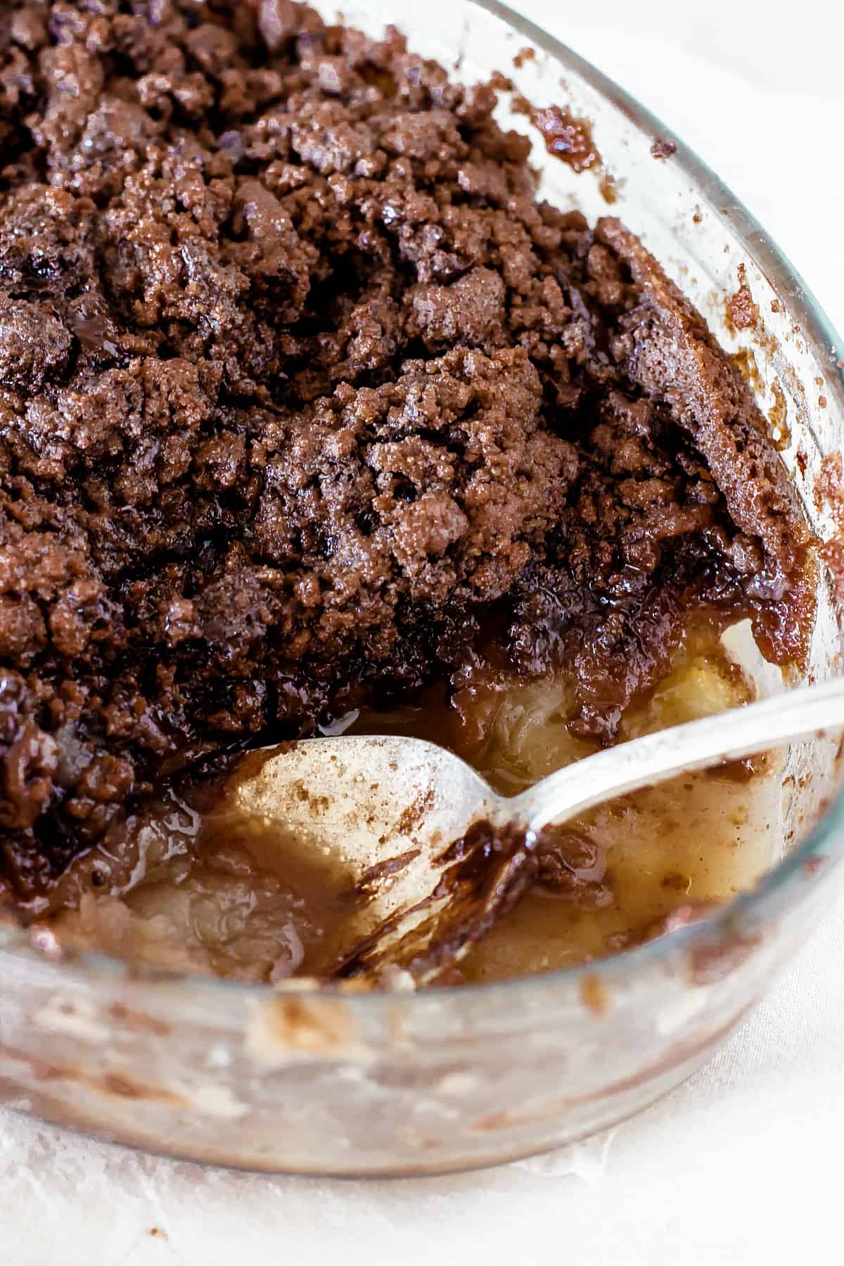 Top view of a silver spoon resting inside glass dish with chocolate apple crisp