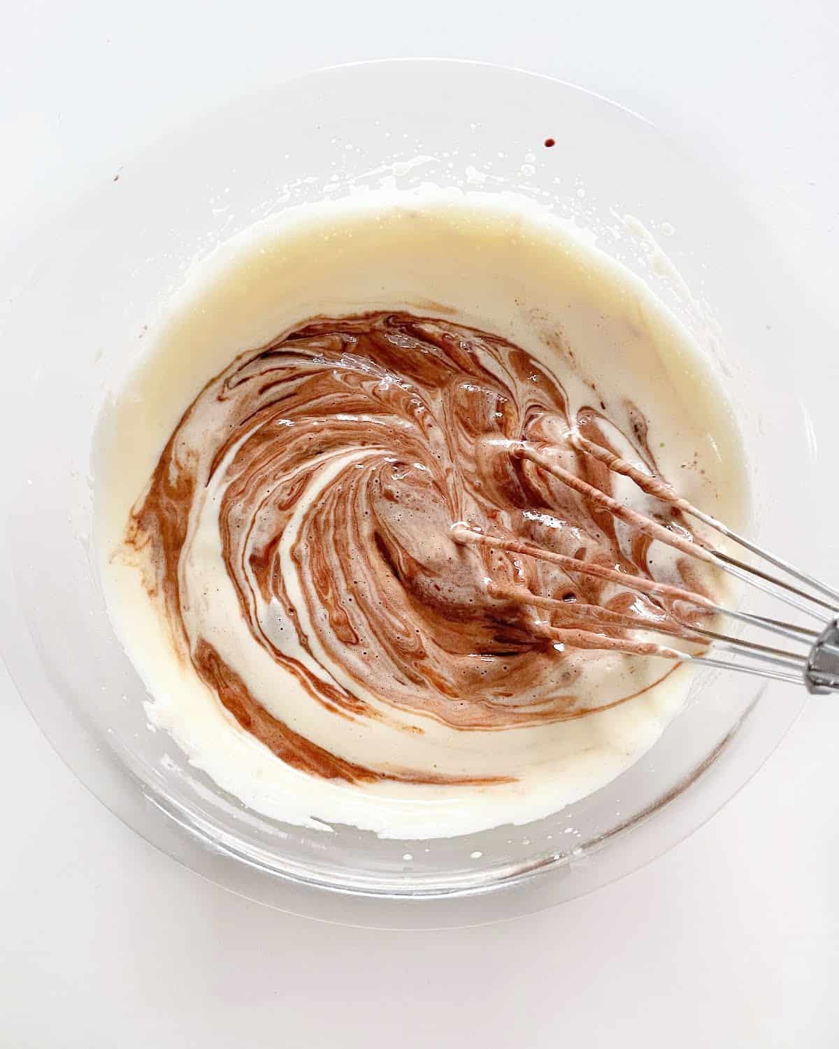 Chocolate and whitish mixture being stirred in a glass bowl with a whisk. White surface.