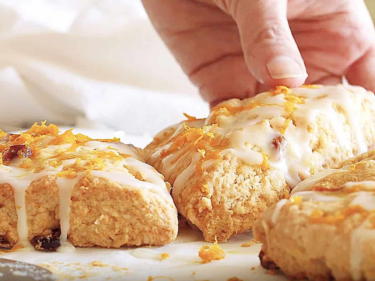 Hand lifting a glazed cranberry orange scone from among others on a white surface.