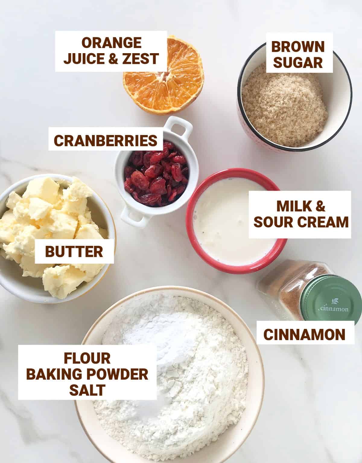 Ingredients for cranberry orange scones in bowls on a white surface including butter, cinnamon, flour mixture, brown sugar, milk and sour cream. .
