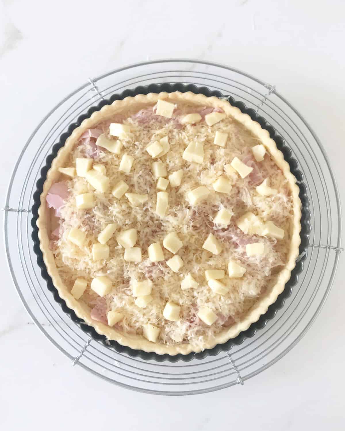 Cheeses over ham layer on pie crust on a wire rack on a white marble surface.