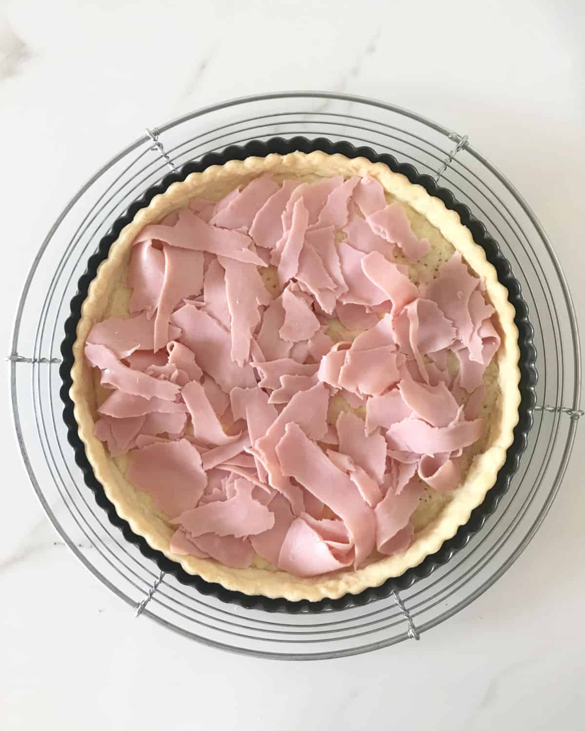 Ham slices on tart crust in dark pan on a metal rack. White marble surface.