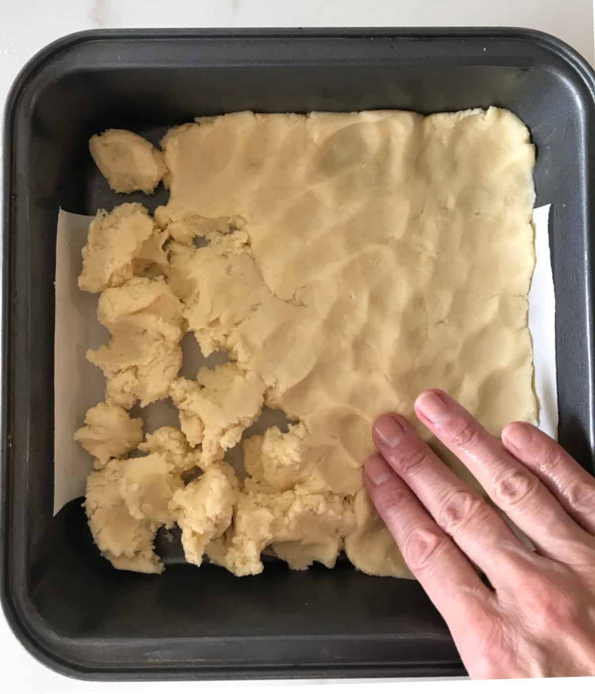 Patting shortbread dough with a hand into a dark metal square baking pan.