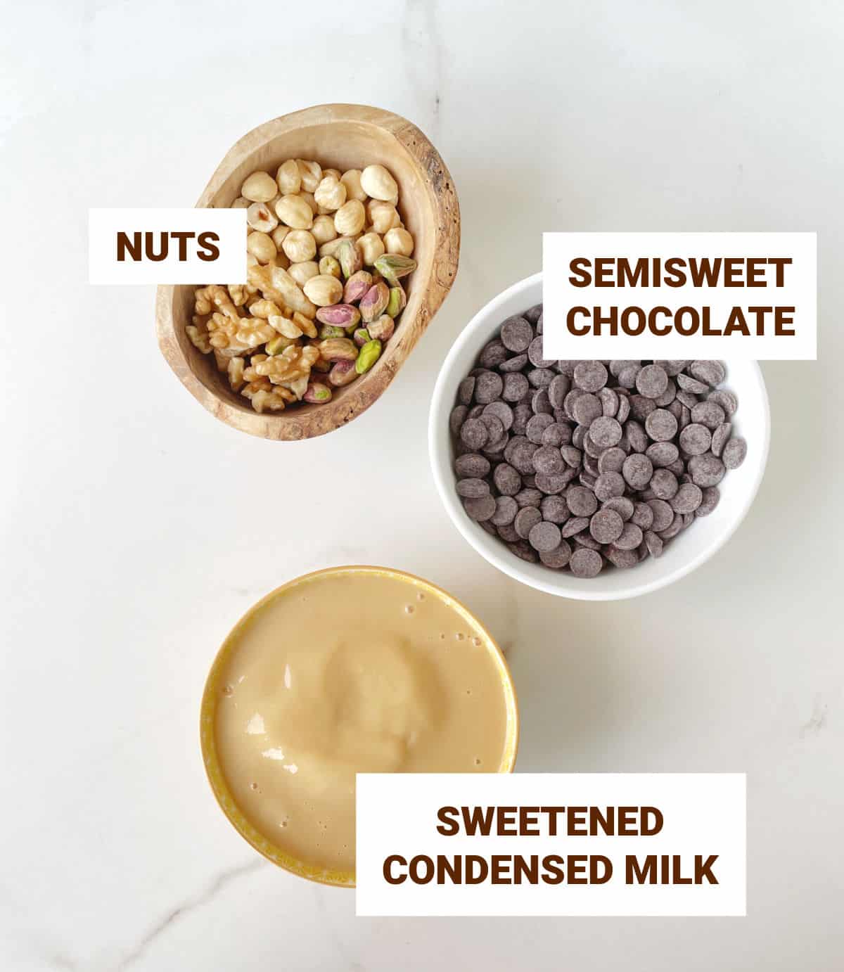 Three bowls on a white marble surface with ingredients for fudge: chocolate chips, condensed milk, and nuts.