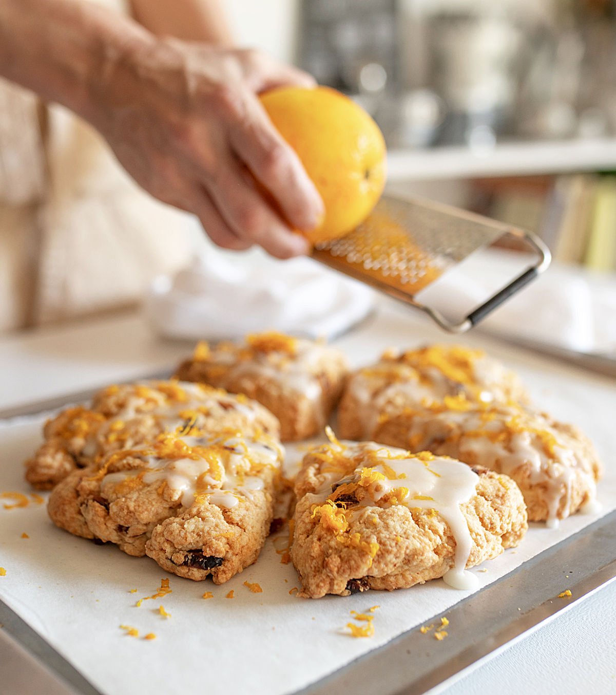 Grating orange peel over glazed scones on white paper and metal sheet pan.